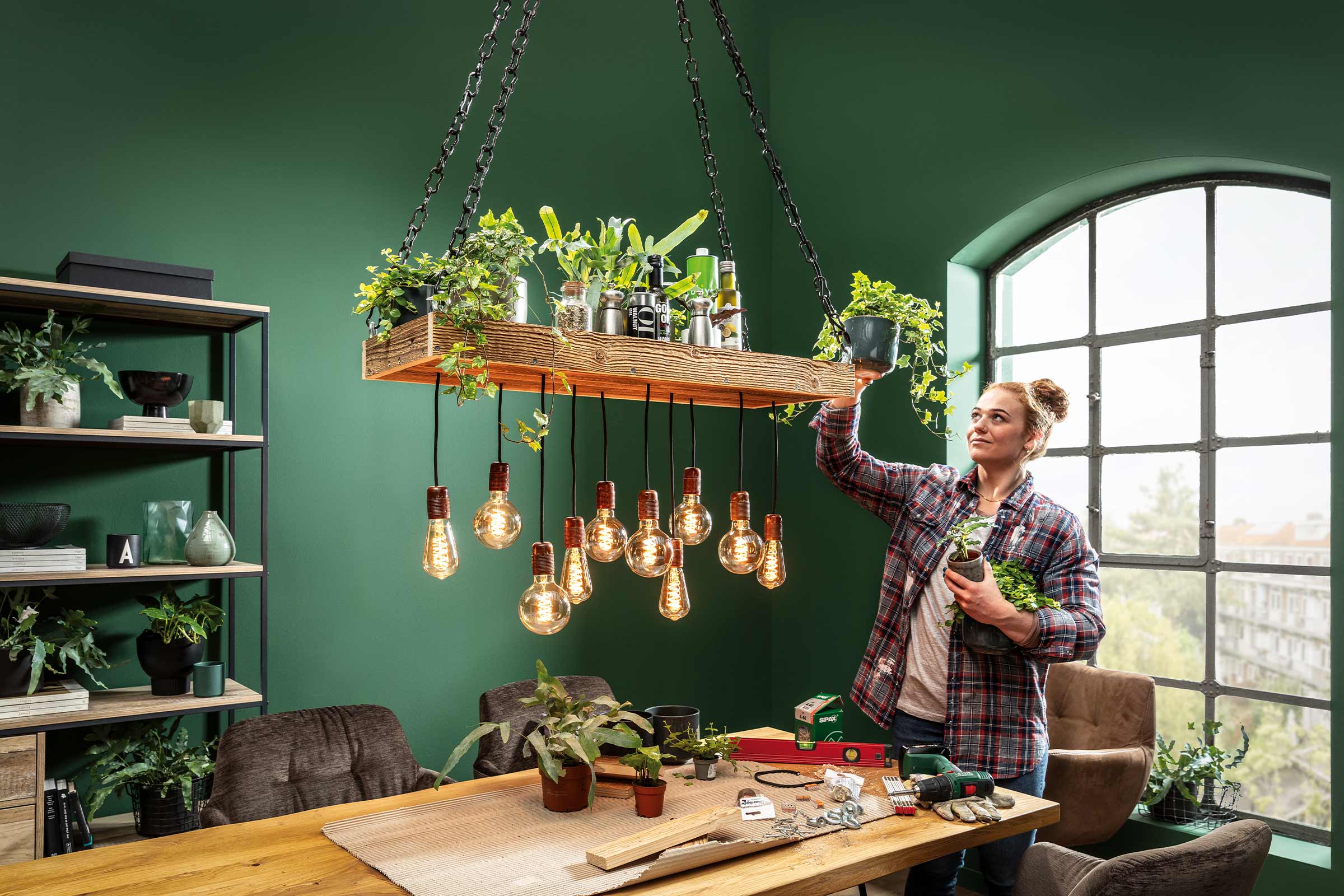 Licht, uitnodigend interieur met een vrouw die kamerplanten in een houten hanglampframe met gloeilampen zet, voor een natuurlijke sfeer.