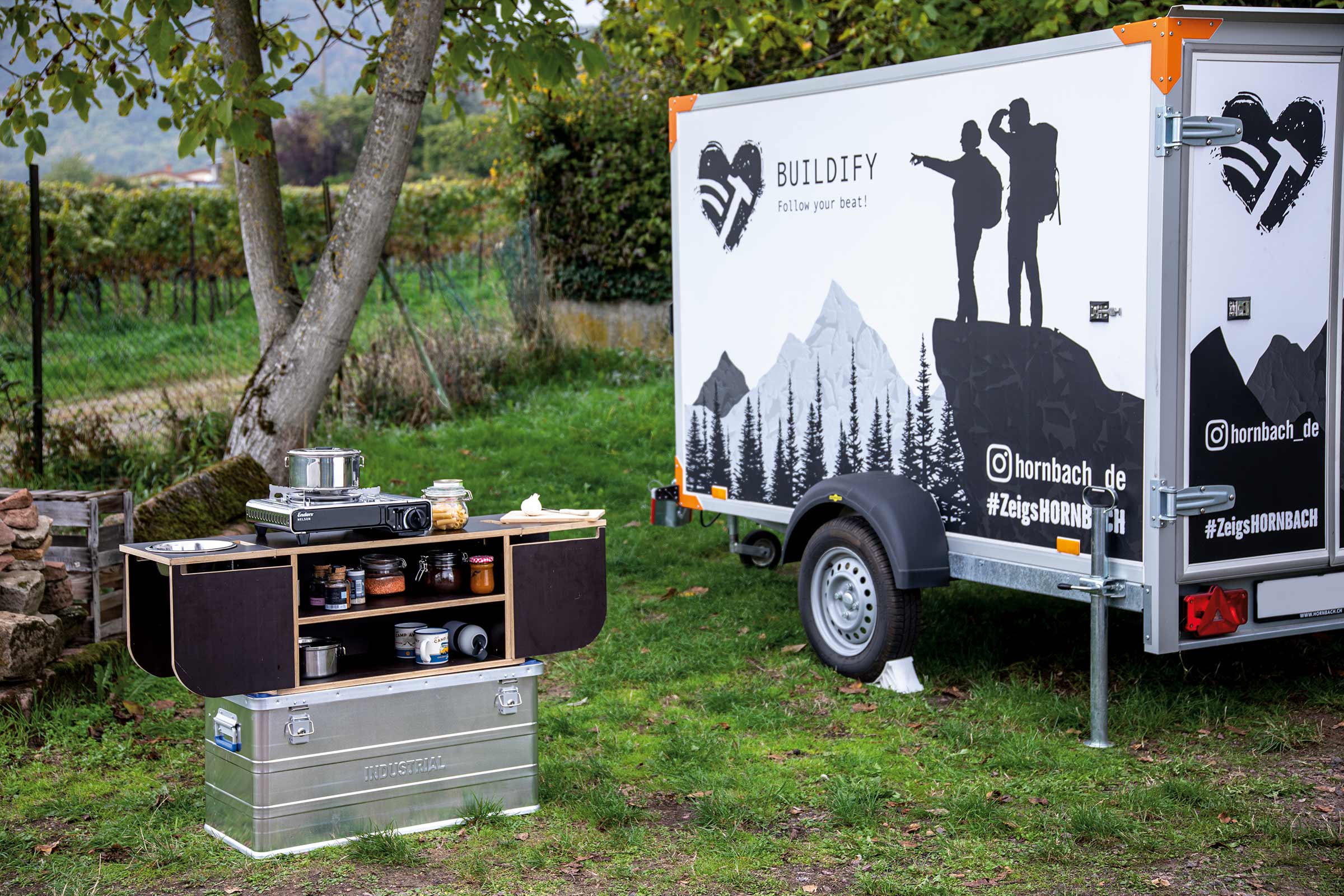 Campingkeuken met kooktoestel, proviand en opbergruimte op een veld met een aanhanger op de achtergrond.