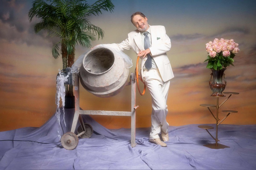 Een man in een pak poseert naast een betonmolen, een bos rozen en een palmboom voor een geschilderde achtergrond.