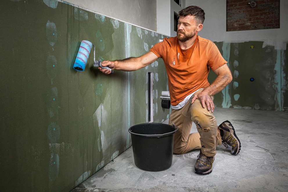 Ambachtsman schildert muur met verfroller in een kamer, emmer staat op de grond