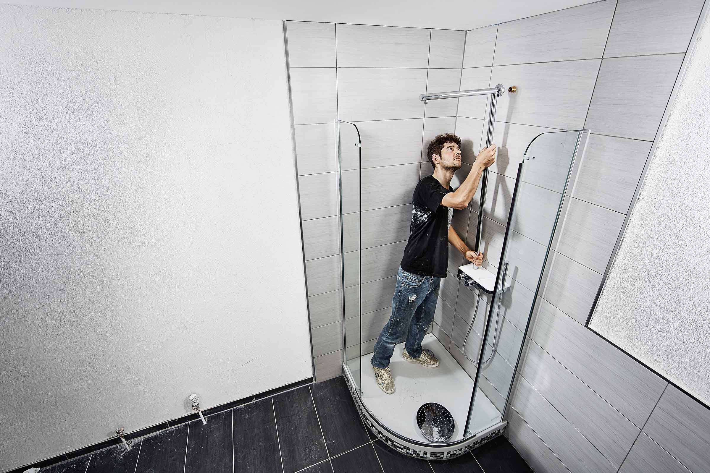 Een man installeert een douchecabine in een badkamer met grijze tegels en een donkere vloer.