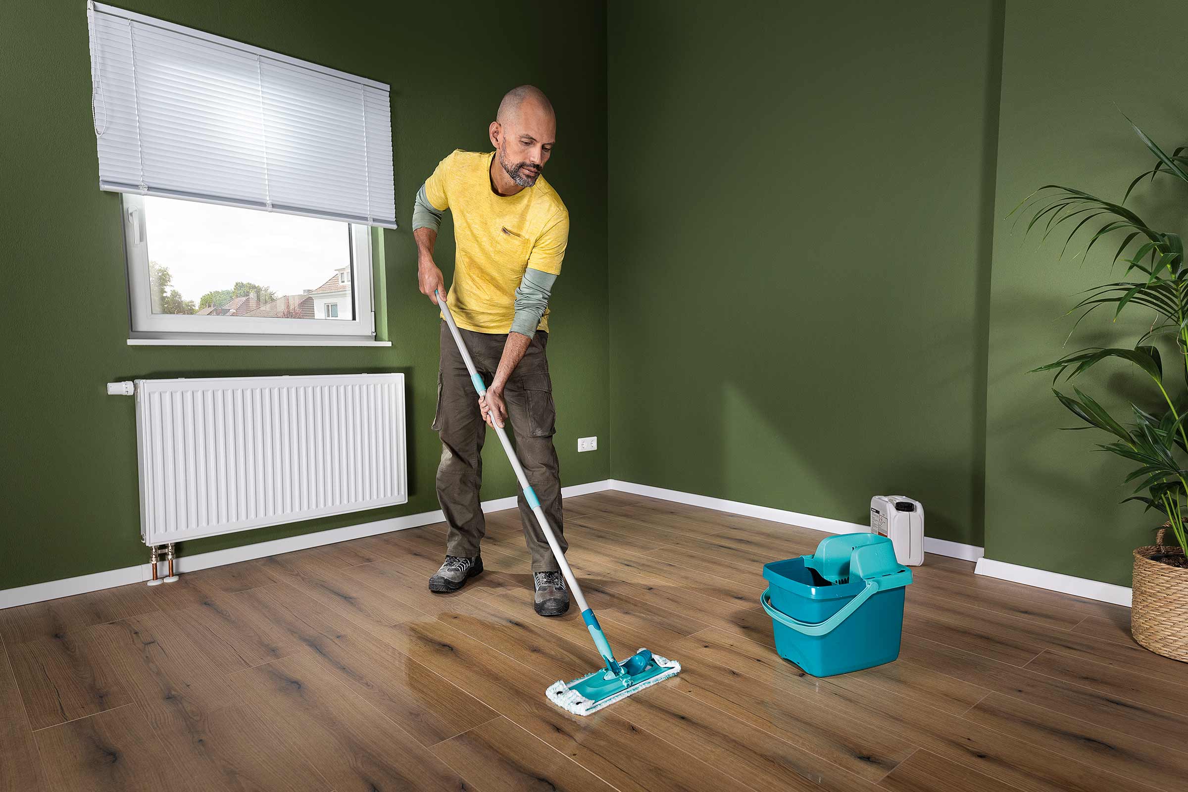 Een man dweilt een houten vloer met een vloerwisser in een kamer met een raam, radiator en potplant