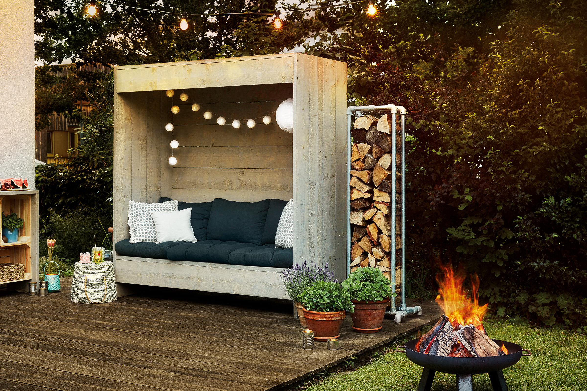 Gezellige tuinscène met houten terras, zithoek met kussens, stapel hout, bloempotten en een kampvuurplaats