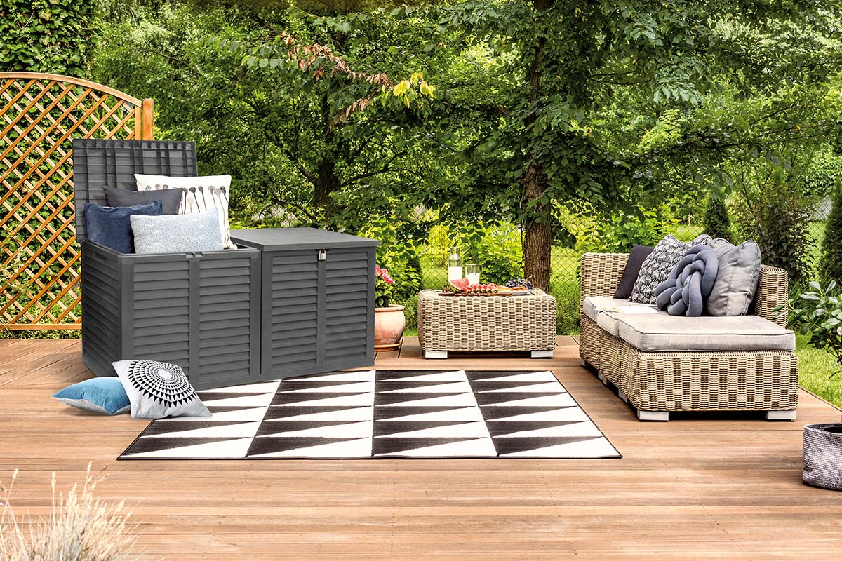 Tuinsetting met grijze opbergbox, rotanmeubels en een geometrisch tapijt op een houten terras met groene achtergrond