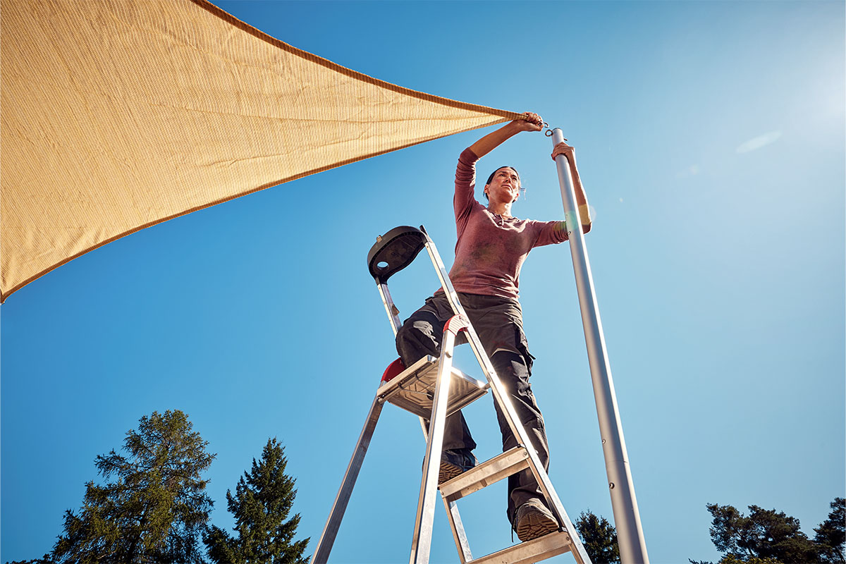 Een vrouw monteert een schaduwdoek op een ladder aan een paal.