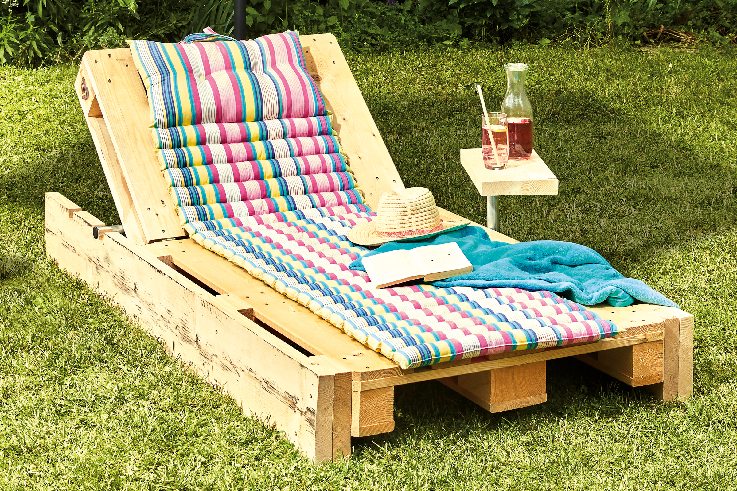 Houten pallet als ligstoel met kussen, strohoed, boek, handdoek en drankjes in de tuin