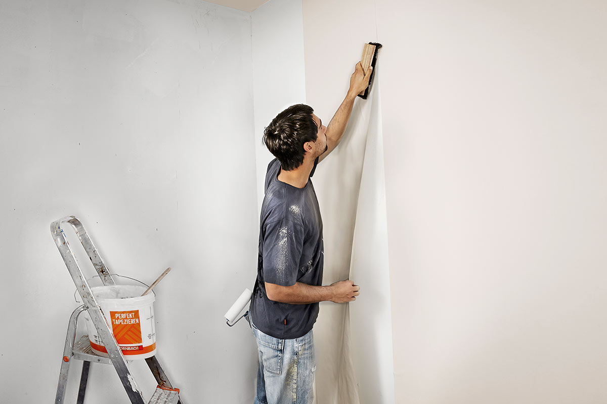Man behangt een muur met een behangborstel, op de achtergrond staan een ladder en een emmer behanglijm.