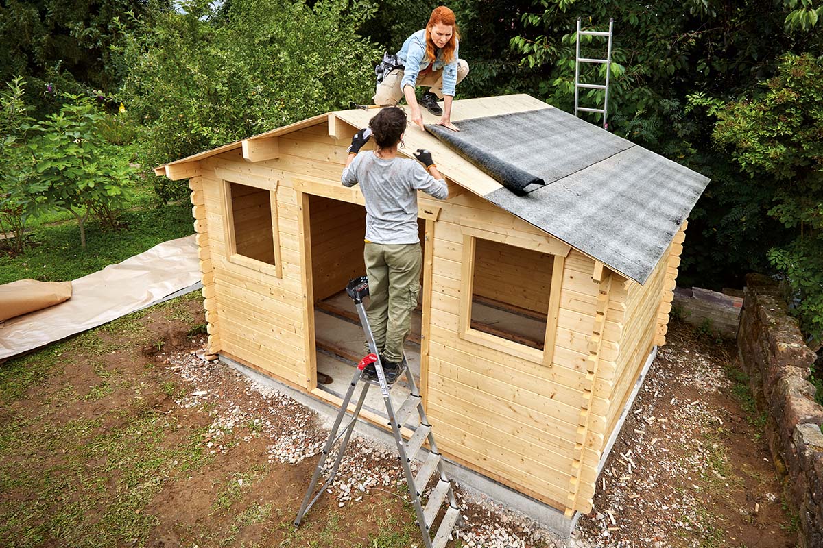 Twee mensen monteren een houten tuinhuis met bitumen dakbedekking op het dak.