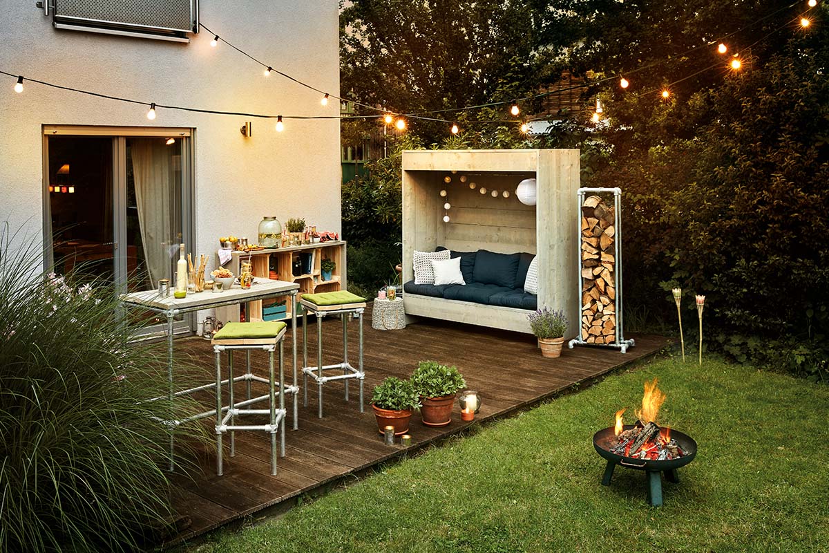 Aangelegde tuin met houten terras, zithoek, statafel, vuurkorf en lichtsnoer voor een gezellige sfeer.