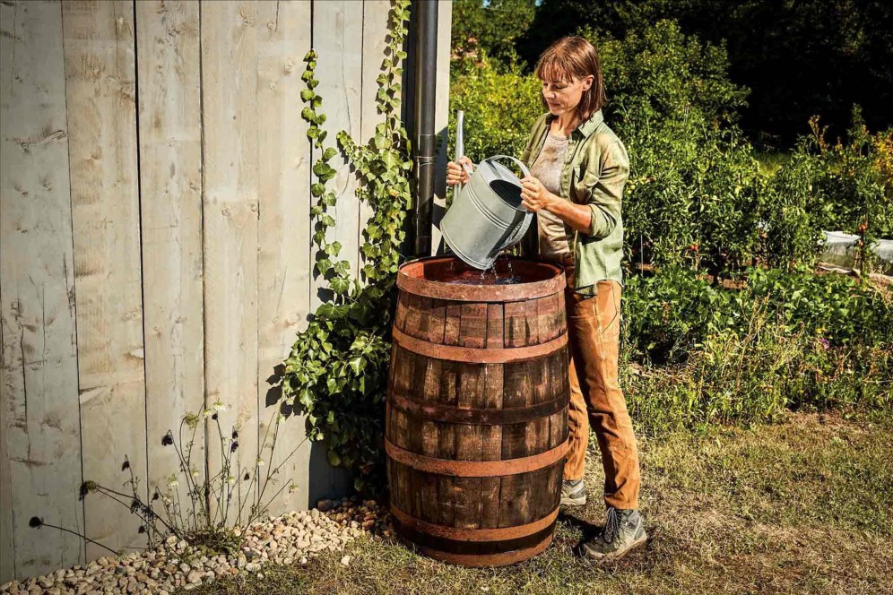 Een vrouw vult een regenton met een gieter in een tuin.