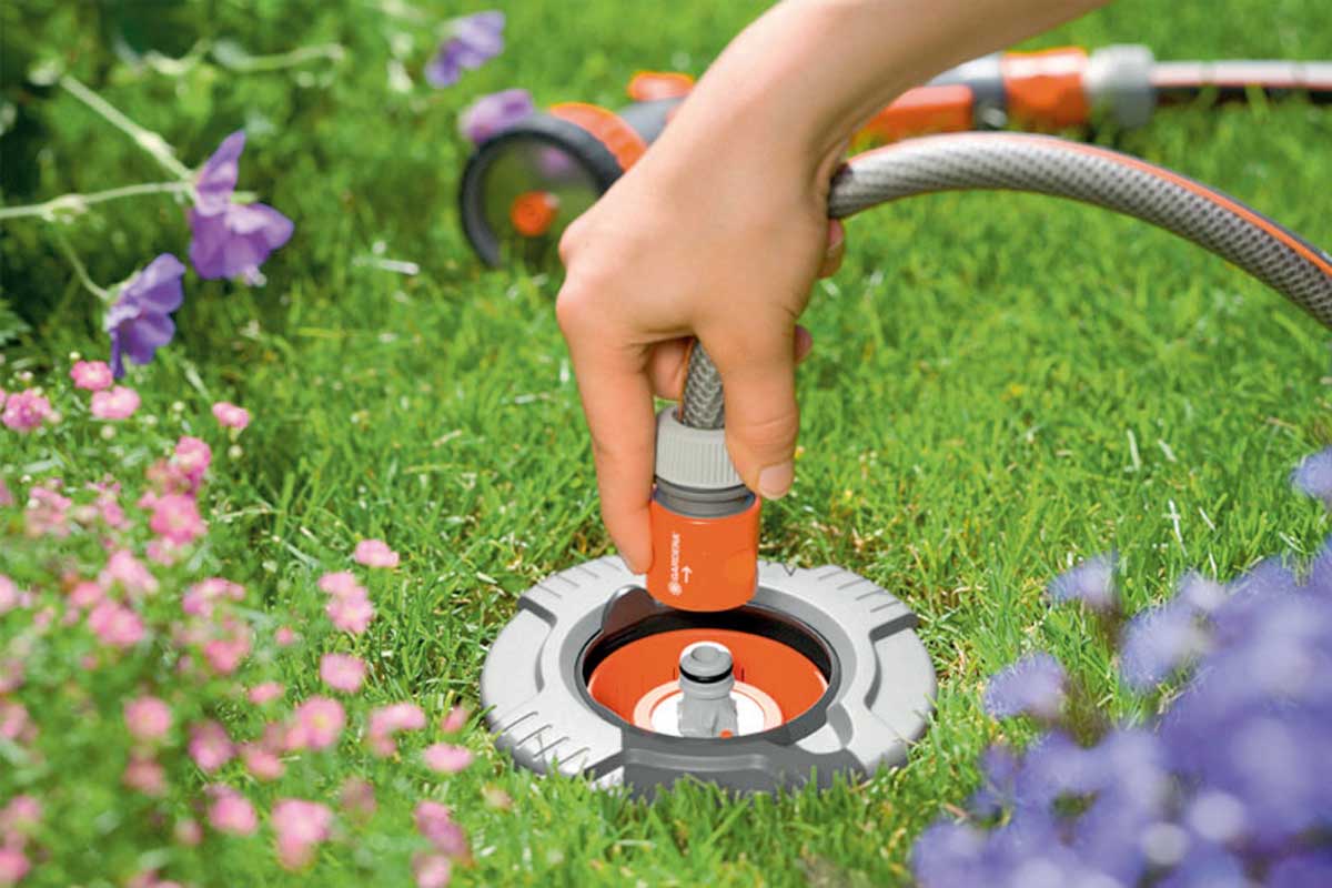 Een hand sluit een tuinslang aan op een ondergrondse wateraansluiting. Tuinslangwagen op de achtergrond.
