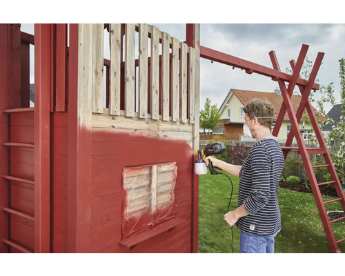 Een persoon spuit een houten speelhuis met een verfspuitpistool