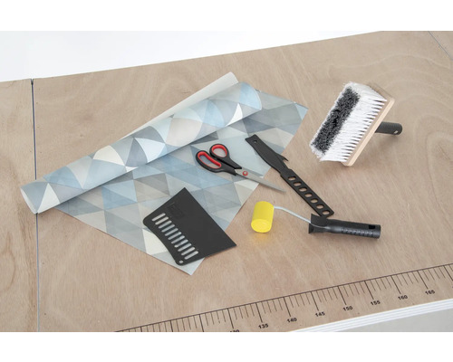 Behang met geometrisch patroon, behangborstel, naadroller, behangmes, aandrukspatel en schaar op een tafel.
