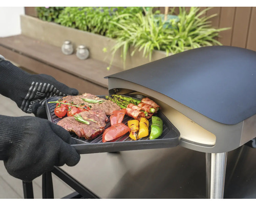 Een persoon met barbecuehandschoenen schuift een grillplaat met steak en groenten in een pizzaoven.