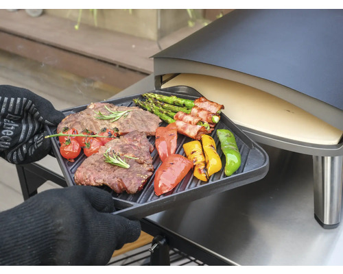 Een persoon met ovenwanten houdt een grillplaat met gegrilde steak, asperges, tomaten en paprika voor een pizzaoven.