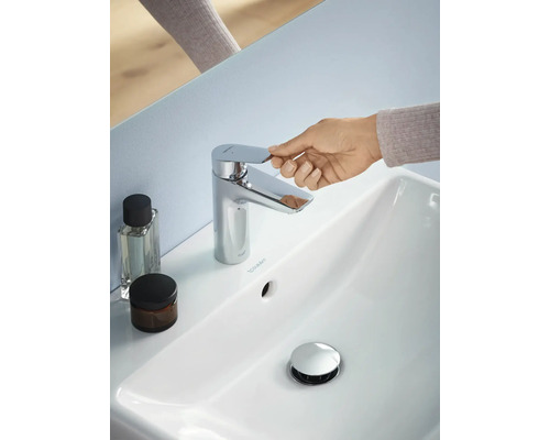 Foto van een badkamer met een witte wastafel, een chromen kraan, een hand die de kraan bedient en twee houders