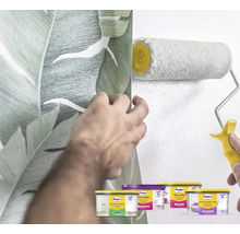 Een man behangt een muur met een behang met plantmotief en schildert een andere muur met een verfroller. Op de voorgrond staan diverse verfcontainers.