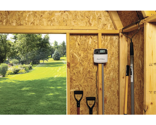 Scene van een tuinirrigatiesysteem met Rain Bird controller en tuingereedschap in een houten schuur, uitzicht op een gazon met actieve irrigatie