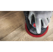 Een hand met een handschoen bedient een excentrische schuurmachine op een houten vloer.