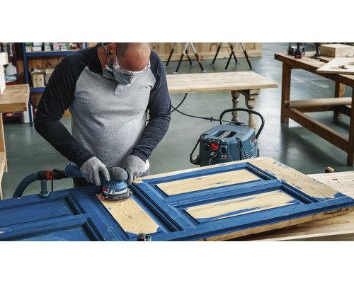 Een persoon schuurt een blauwe houten deur met een excentrische schuurmachine aangesloten op een stofzuiger.