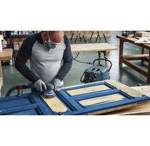 Een persoon schuurt met een excentrische schuurmachine een blauwe houten deur, ondersteund door een stofzuiger.