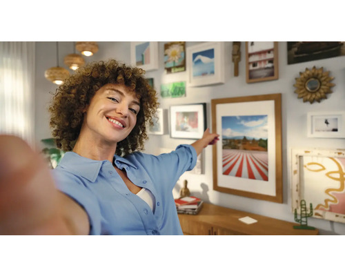 Vrouw wijst naar een muur met ingelijste foto''s en decoratie in een heldere woonkamer.