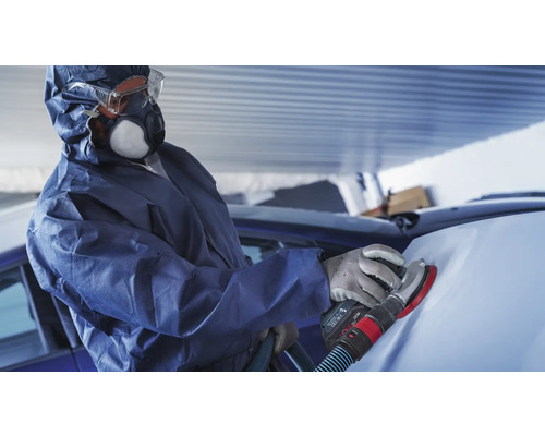 Een man in beschermende kleding schuurt een auto met een schuurmachine.