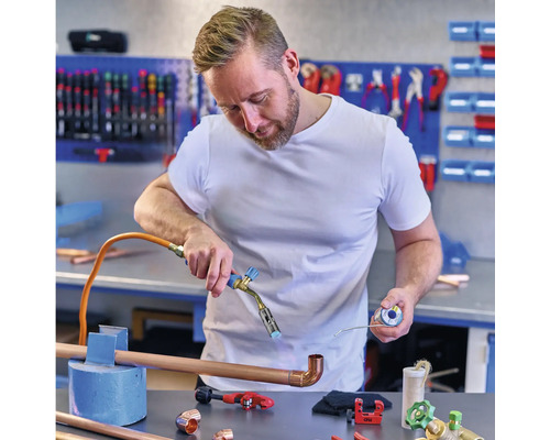 Een man soldeert koperen buizen met een soldeerbout in een werkplaats, met koperen fittingen en gereedschap op de tafel.