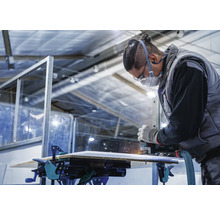 Een vakman freest een plaat met een bovenfrees, die met lijmklemmen op een werkbank is bevestigd.