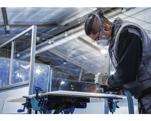 Een vakman freest een plaat met een bovenfrees, die met lijmklemmen op een werkbank is bevestigd.