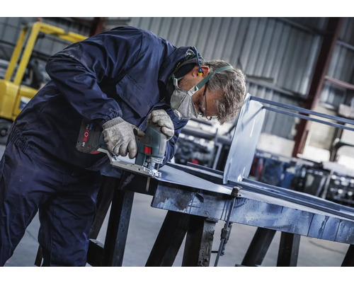 Man met veiligheidsmasker, handschoenen en gehoorbescherming zaagt een stuk metaal met een decoupeerzaag op batterijen