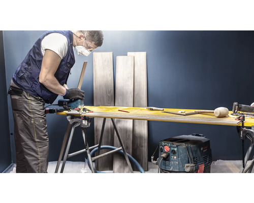 Vakman snijdt laminaat met een decoupeerzaag op een werkbank. Op de achtergrond staan laminaatplanken.