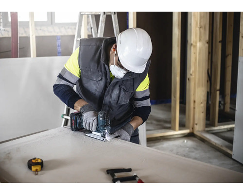 Een vakman zaagt een gipsplaat met een decoupeerzaag terwijl hij beschermende kleding draagt.