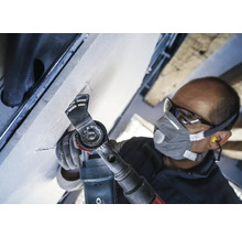 Ambachtsman aan het werk met een multifunctioneel gereedschap aan een plafond, uitgerust met een veiligheidsbril en een stofmasker