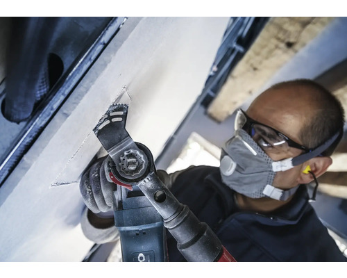 Ambachtsman aan het werk met een multifunctioneel gereedschap aan een plafond, uitgerust met een veiligheidsbril en een stofmasker