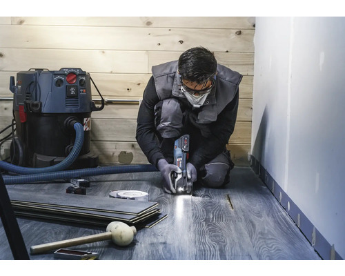 Een man knielt en snijdt laminaat met een multifunctioneel gereedschap. Een stofzuiger, laminaatvloeren en gereedschap zijn ook zichtbaar.