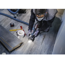 Een vakman snijdt laminaat met een multifunctionele gereedschapsmachine om een uitsparing voor een pijp te creëren. Gereedschappen zoals een meetlint en een potlood liggen klaar.