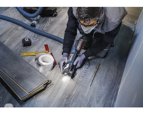Een vakman snijdt laminaat met een multifunctionele gereedschapsmachine om een uitsparing voor een pijp te creëren. Gereedschappen zoals een meetlint en een potlood liggen klaar.