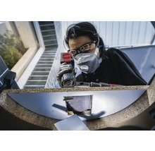 Vrouw met veiligheidsbril en stofmasker werkt met een multislijper aan een werkstuk