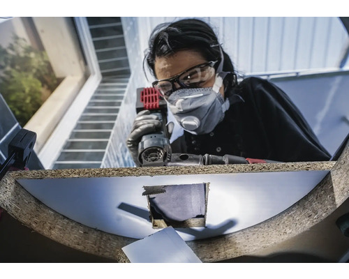 Vrouw met veiligheidsbril en stofmasker werkt met een multislijper aan een werkstuk