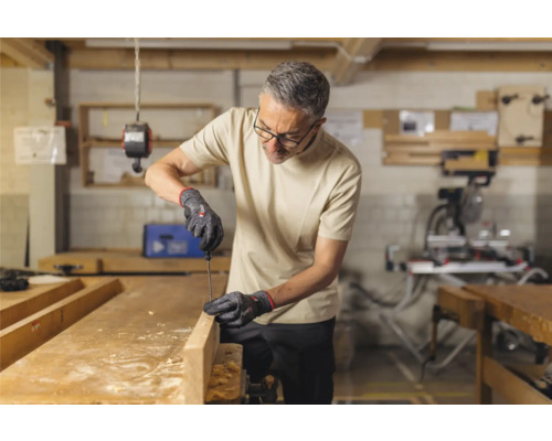 Man werkt met schroevendraaier en handschoenen aan een houten plank in een werkplaats.