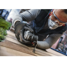 Een man met een veiligheidsbril, ademhalingsbescherming en handschoenen werkt met een multifunctioneel gereedschap aan een houten plank.