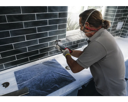 Tegelzetter aan het werk met een multifunctioneel gereedschap op een wand met zwarte tegels.