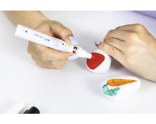 Iemand beschildert een steen met een rode stift, terwijl een andere steen met een wortel op de voorgrond ligt.