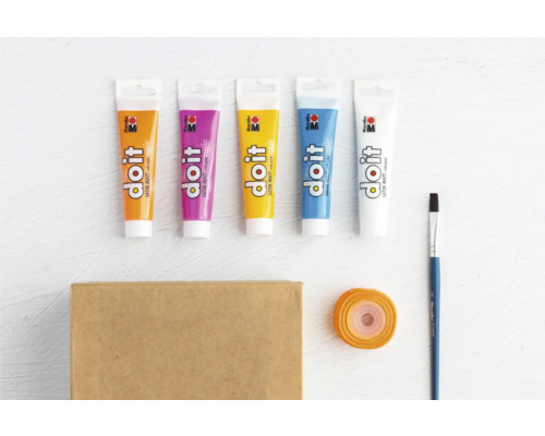 Diverse tubes verf, penseel, cadeaupapier en een kartonnen doos liggen op een tafel.