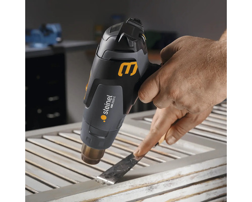 Een persoon verwijdert verf van een raamkozijn met een heteluchtpistool en een plamuurmes.