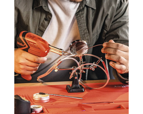 Iemand soldeert draden met een soldeerpistool, die zijn bevestigd met een houder met een vergrootglas. Op het werkoppervlak bevinden zich fluxmiddel en reinigingsaccessoires.