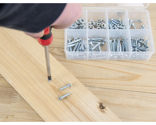 Iemand schroeft een schroef met een schroevendraaier in een houten plank, naast een assortimentsdoos met schroeven.