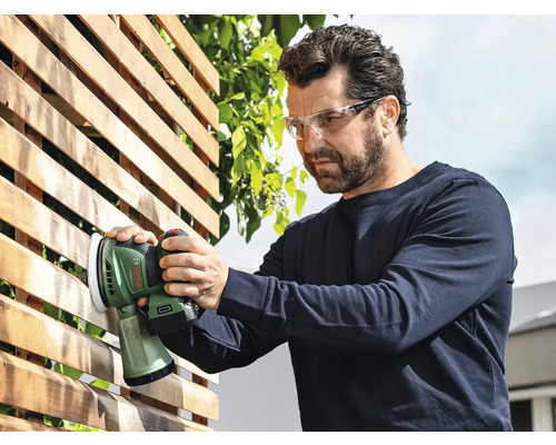 Man schuurt een houten schutting met een Bosch accu-excentrische schuurmachine en draagt een veiligheidsbril.