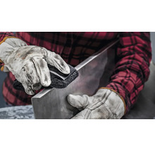 Een persoon reinigt een stuk metaal met een schuurborstel, beschermd door werkhandschoenen en een geruit overhemd.